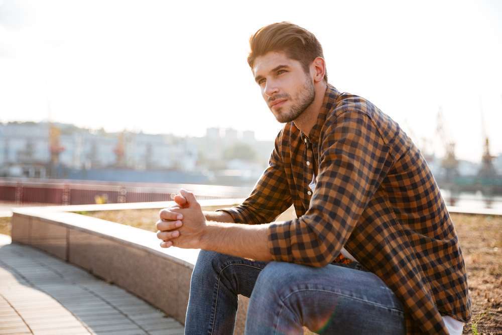 Pensive young man in plaid shirt sitting and thinking outdoors